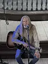En mann med langt hår sitter med gitar foran en mikrofon i et kirkerom.
