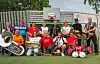 Gruppe som poserer utendørs med fotballer, flagg og et stort blåseinstrument foran en basketballkurv.