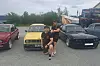 To personer står foran en gul og en svart bil på en parkeringsplass med andre kjøretøy bak.