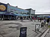 Uteområde foran et moderne bygg med folk, skilt og en blå markise ved inngangen.