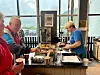 En kvinne serverer mat ved en buffetdisk i et lyst lokale mens en blid mann forsyner seg med kake.