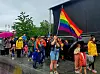 Personer går i parade med regnbueflagg ved en stor scene på en våt dag.