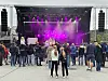 Folk står foran en utendørs konsertscene med lilla scenelys. To på scenen.