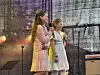 To unge står på en scene med mikrofoner. Lys og musikkutstyr i bakgrunnen.