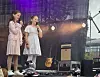 To unge står på en scene med mikrofoner. Lys og musikkinstrumenter i bakgrunnen.