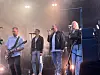 Et band spiller live på en scene med mikrofoner, gitar og sterke lys.