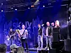Band spiller på scene med mikrofoner, gitarer og blå scenebelysning.