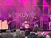 Et band spiller på en scene med Skuv-bakgrunn og publikum i forgrunnen.