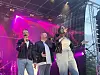 Tre artister synger og opptrer på en utendørsscene med rosa scenelys.