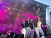 Artister synger på en utendørsscene med rosa lys og publikum foran.