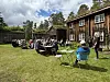 Gruppe med folk sitter ved langbord og campingstoler på en gressplen foran et rødt trehus og musebygninger.