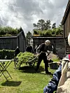 Frivillige på et gresslagt tun med trillebår, stoler og gamle trebygninger i bakgrunnen.