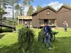 To personer bærer store bunter med gras på en grønn plen foran trebygninger. hesjing