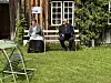 To personer sitter på benker foran en trebygning med vinduer, omgitt av gress, en stol og grønn beplantning.