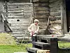 En gutt står ved et gammelt trehus med benker, redskaper og et bord på graset.