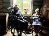 To kvinner sitter i en mørk, tradisjonell stue ved et vindu med røde blomster i potte.