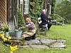 En person sitter ved en bøtte med blomster ved en trebygning, mens en annen sitter på en benk på gresset.