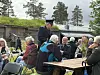 Folk sitter rundt bord og i stoler på et uteområde med trær, bygninger og servering.