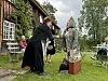 Folk står og sitter på et gresskledd tun ved en trebygning med vinduer, bord og stoler i bakgrunnen.