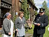 Tre personer står utenfor et tømmerbygg, mens en uniformert mann leser fra papir ved inngangen.