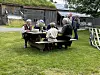 Flere personer sitter og står rundt et rundt trebord på gresset ved et gammelt bygg.