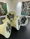 Abstrakt skulptur med svarte mønstre i et gallerirom med to store kunstverk på veggen.