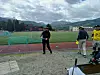Flere personer med diskgolfskiver står ved en løpebane og fotballbane med fjell og overskyet himmel i bakgrunnen.