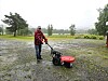 Person som går bak en liten maskin på en våt grusplass under overskyet himmel.