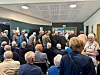 Tettpakket folkemøte i et møterom med folk som står og sitter foran en skjerm.