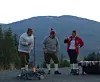 Tre personer står rundt et lite bål foran en fjellside og trær.