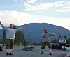 Tre personer står og danser på en grusplass med fjell og trær bak.