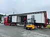 Rød lastebil med åpen semitrailer og flere containere ved en industriell vei, med en truck foran.