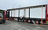 Stor semitrailer festet med stropper på en lastebiltransporter, med to personer ved siden av.