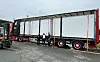 Stor semitrailer festet med stropper på en lastebiltransporter, med to personer ved siden av.