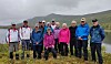 En gruppe på ti personer står samlet ute i naturen med fjell og et vatn bak.