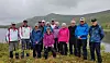 En gruppe på ti personer står samlet ute i naturen med fjell og et vatn bak.