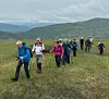 En gruppe turgåere går over en grasbakke med fjell og skyer i bakgrunnen.
