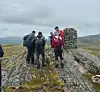 Fire turgåere står ved en steinvarde på en fjelltopp under en grå, overskyet himmel.