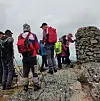 Turgåere med ryggsekker står på en steinete fjelltopp ved en steinrøys i overskyet vær.