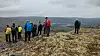 Flere turgåere står på en steinete fjellrygg med utsikt over dal og vann i grått vær.