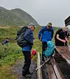 Flere turgåere med ryggsekker og regnjakker står ved en tretrapp i et fjellandskap.