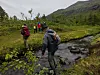 Flere vandrere krysser en bekk i et grønt fjellområde med bratte åssider i bakgrunnen.