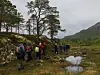 Turgåere med ryggsekker følger en sti ved et vannspeil i et kupert, grønt landskap.