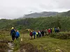 En gruppe vandrere går på en smal sti i et grønt fjellområde med tåke og skyer i bakgrunnen.