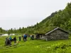 Vandrere står ved en steinhytte i grønt fjellterreng med innsjø og skogkledd ås.
