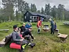 Flere personer sitter og står samlet utenfor en hytte med bord, benker og flasker på gresset.