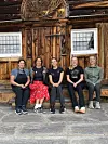 Fem kvinner sitter på en benk foran en brun tømmerbygning med utskårne ornamenter og små vinduer.