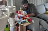 Et barn står ved et lekekjøkken med matleker i en stue, mens en voksen sitter i sofaen.