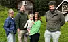 Fire voksne og et lite barn står sammen foran en trehytte på landet.