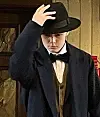 Mann i mørke klær og svart hatt foran en vegg med trepanel.